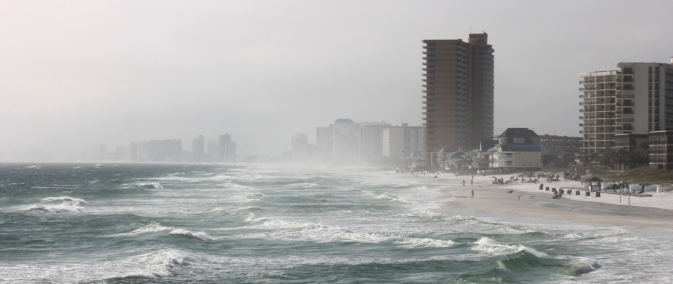  Beach-goers are enjoying the surf on a beach with large waves crashing on shore and buildings in the background while a thick fog moves in unexpectedly.